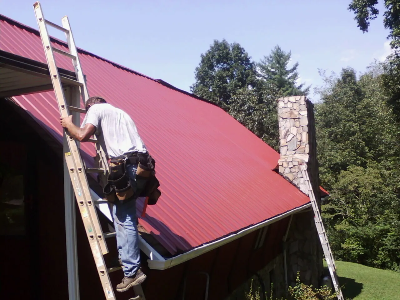 Roofer on a ladder beside a red metal roof and stone chimney — Soffit & Fascia Repair by Roofing Brothers in Floral